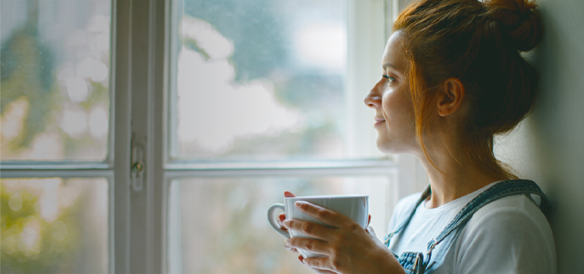 Frau sitzt gemütlich mit einer Tasse Tee in der Hand vor einem Fenster und schaut hinaus – Symbolbild für eine Glasversicherung