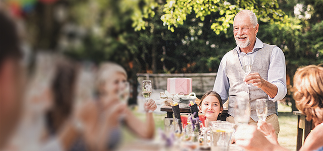 Ein älterer Mann hält bei einem Fest eine Rede an seine Familie, die am Tisch vor ihm sitzt und sie prosten sich mit Sekt zu – Symbolbild für Bestattungsvorsorge